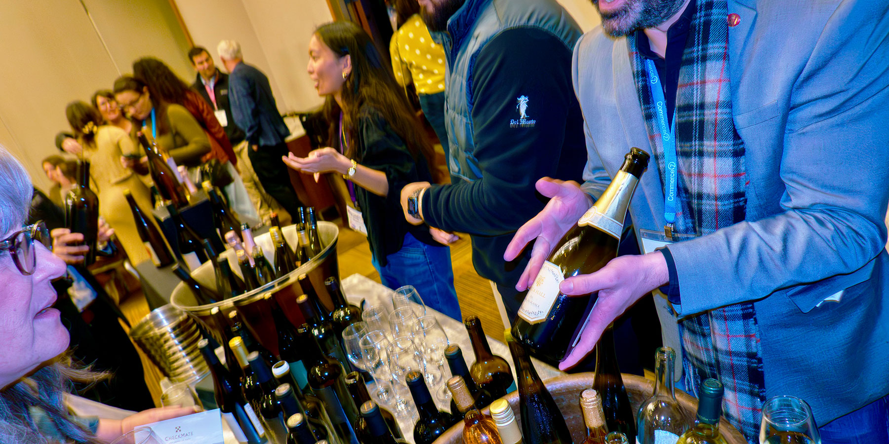People sampling and serving wine at a busy indoor wine tasting event, reminiscent of the vibrant atmosphere at the Direct to Consumer Wine Symposium, with bottles and glasses arranged on tables.