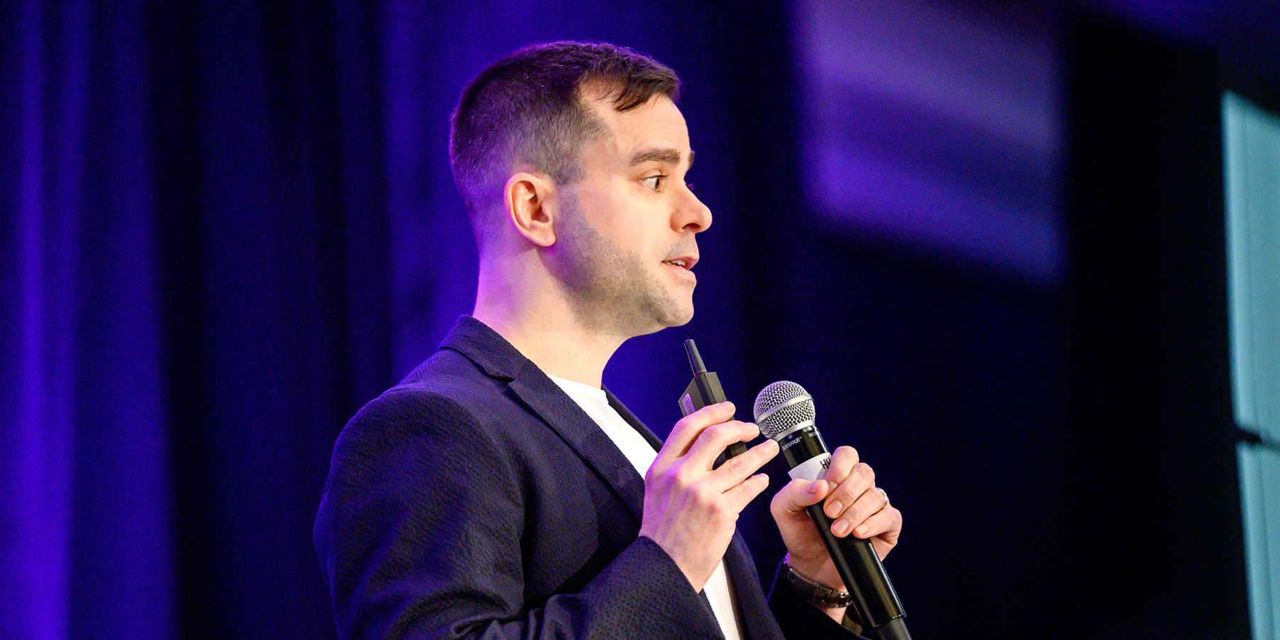 A man in a suit holds a microphone and remote, speaking on stage with a purple background at the Direct to Consumer Wine Symposium.