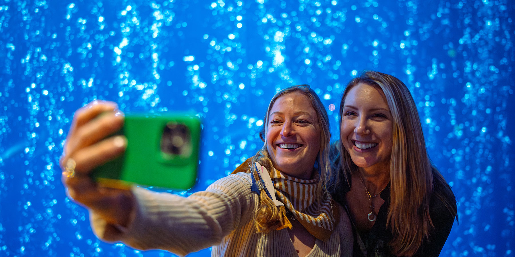 Two women smile and take a selfie together in front of a sparkling blue background at the Direct to Consumer Wine Symposium.