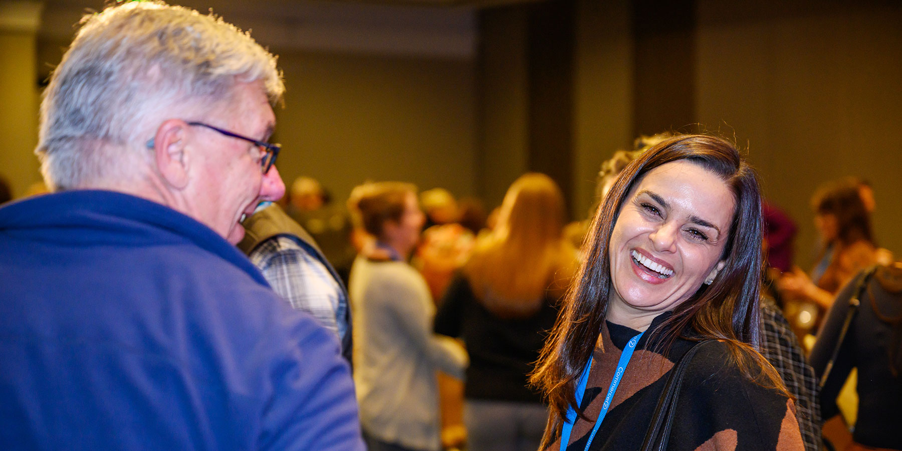Woman smiling and laughing at the Direct to Consumer Wine Symposium, surrounded by other people in the background at this lively indoor social event.
