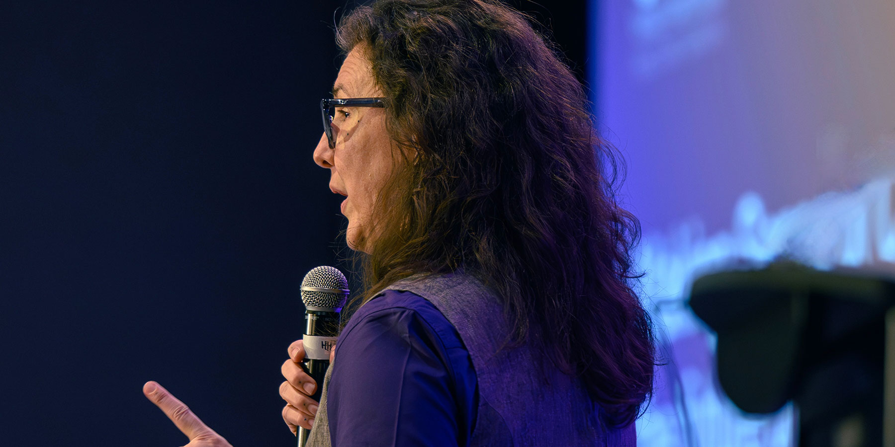Person with long hair and glasses speaking into a microphone on stage at the Direct to Consumer Wine Symposium, with a blurred background.