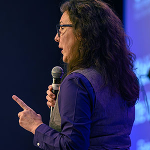 Person with long hair and glasses speaking into a microphone, gesturing with one finger raised as they talk about an important point.
