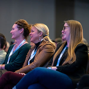 Three women sit in an audience, smiling and laughing about something amusing, wearing conference lanyards around their necks.