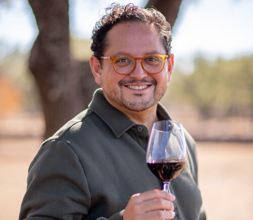 Man with glasses smiling and holding a glass of red wine outdoors, with trees in the background.
