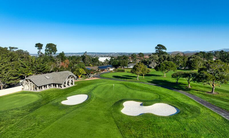 A golf course with sand bunkers, green fairways, trees, and a clubhouse under a clear blue sky.