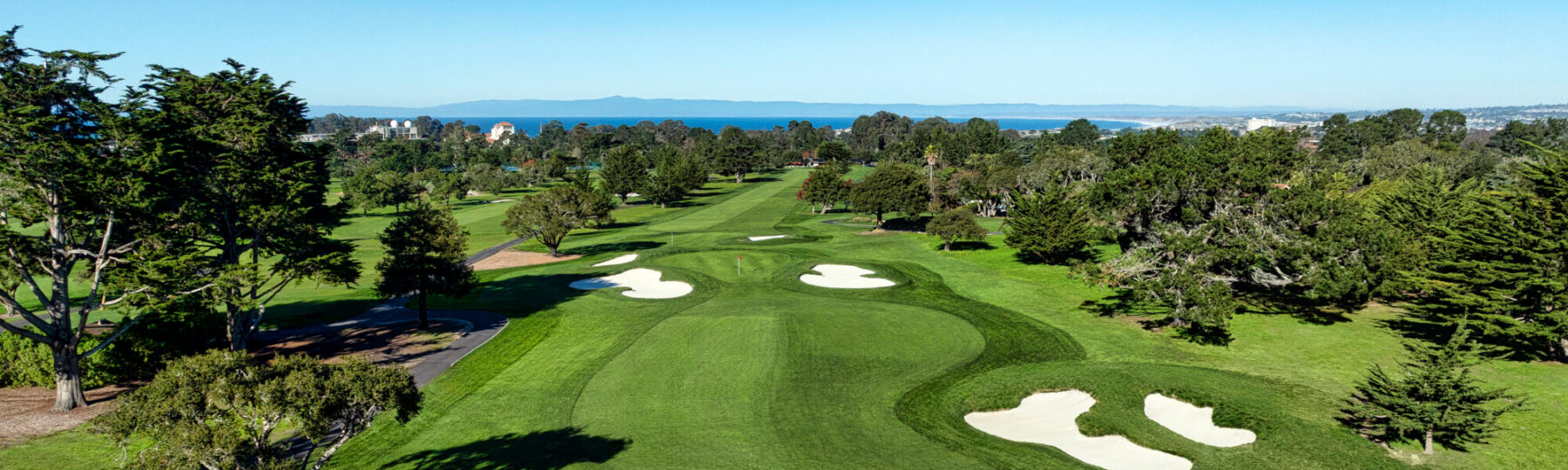 Aerial view of a green golf course with sand bunkers, trees, and ocean in the background under a clear sky.