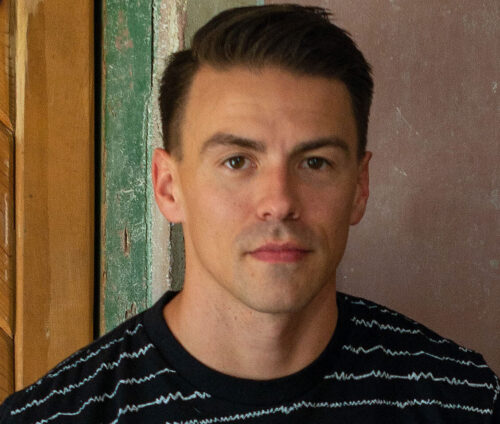 A man with short dark hair and a black patterned shirt stands indoors near a green and beige wall.