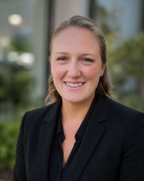 Woman in a black blazer smiling outdoors with greenery and a blurred building in the background.