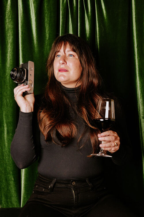 Woman in black holding a vintage camera and a glass of red wine, seated in front of green velvet curtains.
