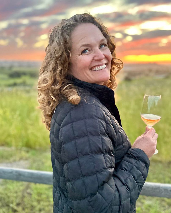 Woman with curly hair in a black jacket smiles while holding a wine glass outdoors at sunset.