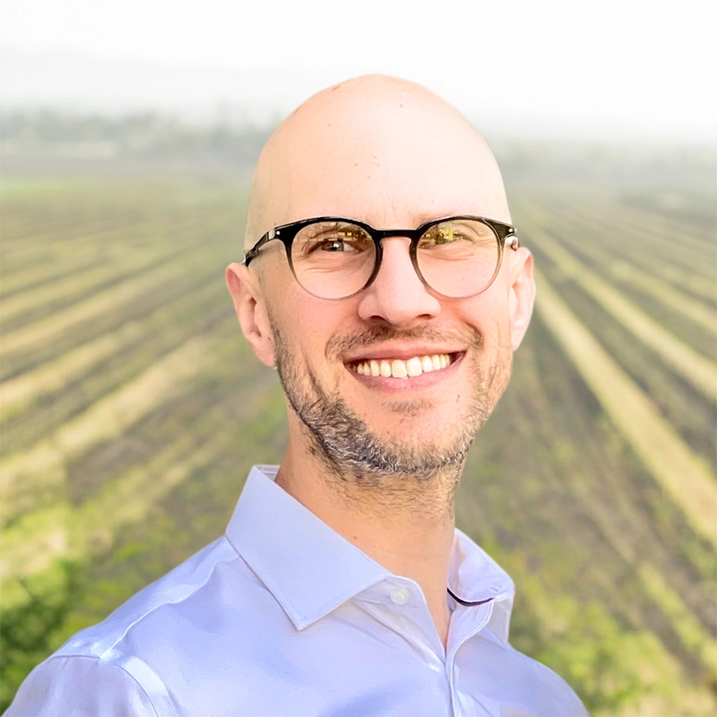 Smiling bald man with glasses standing in front of a blurred, sunlit field with rows of crops.