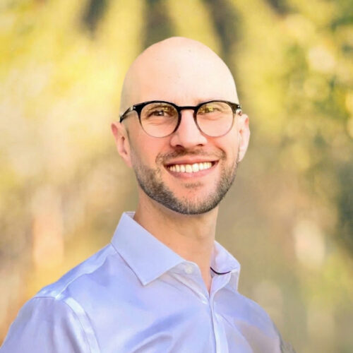 Bald man with glasses smiling, wearing a white shirt, standing outdoors with a blurred green background.