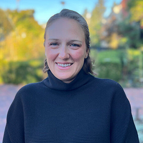 Woman with light brown hair smiling, wearing a black turtleneck sweater, outdoors with greenery in the background.