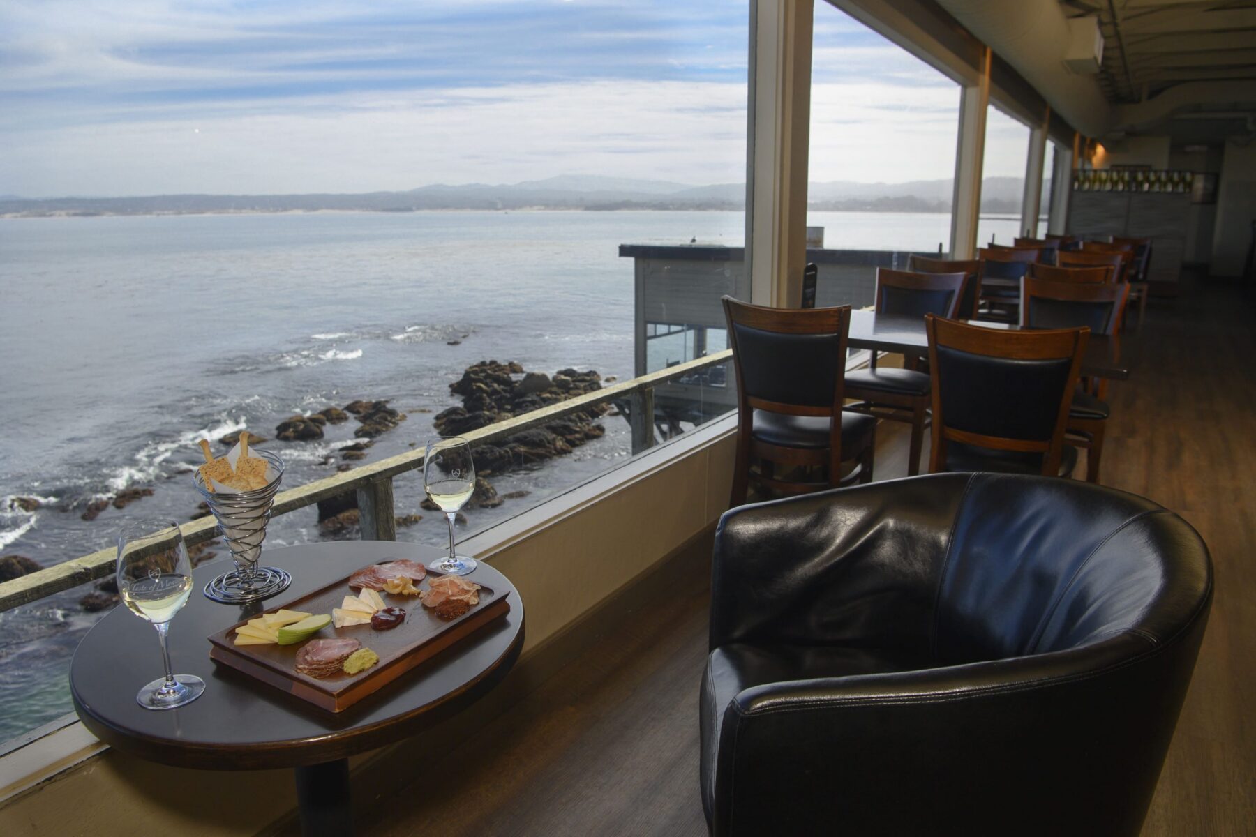 A charcuterie board and two glasses of wine on a table by a window overlooking the ocean.