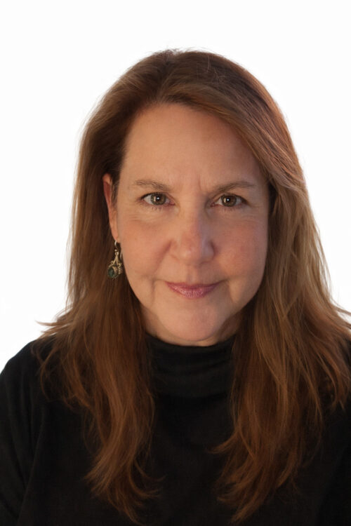 Woman with long, light brown hair and earrings, wearing a black top, smiling slightly at the camera.