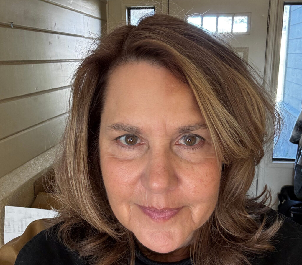 A woman with shoulder-length brown hair smiles slightly, standing indoors near a window and wooden wall.