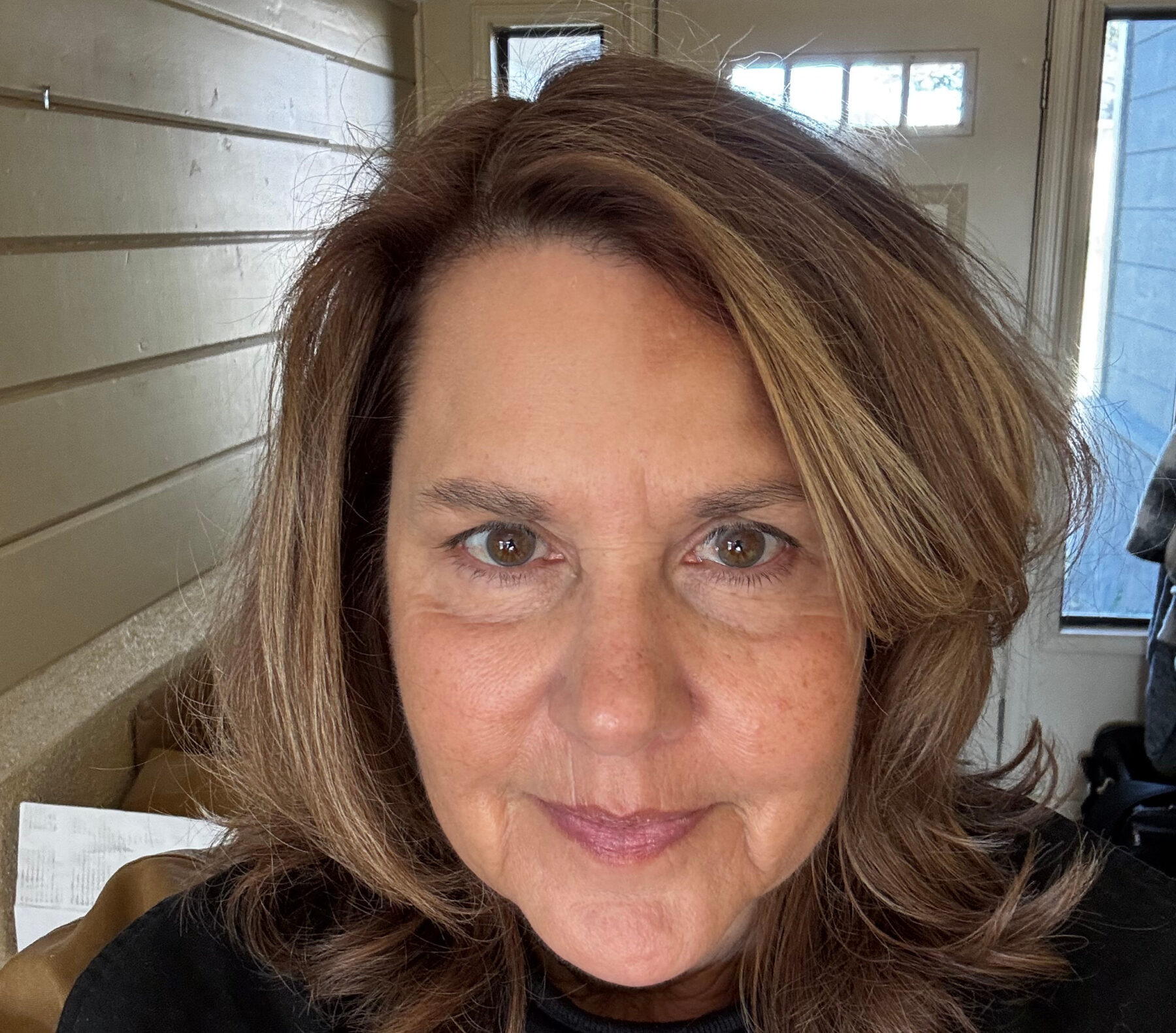 A woman with shoulder-length brown hair smiles slightly, standing indoors near a window and wooden wall.