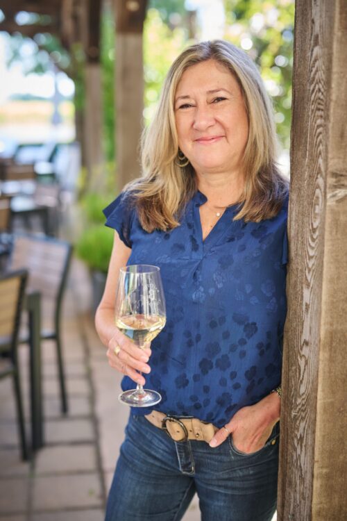 Woman with blonde hair in blue blouse holding a glass of white wine, smiling outdoors by a wooden post.