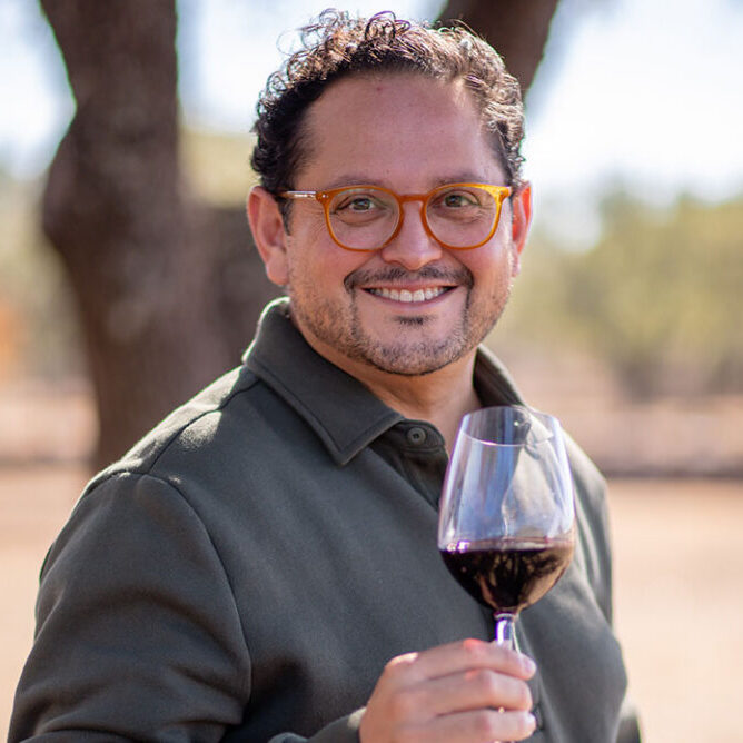 Man with glasses smiling and holding a glass of red wine outdoors, with trees in the background.