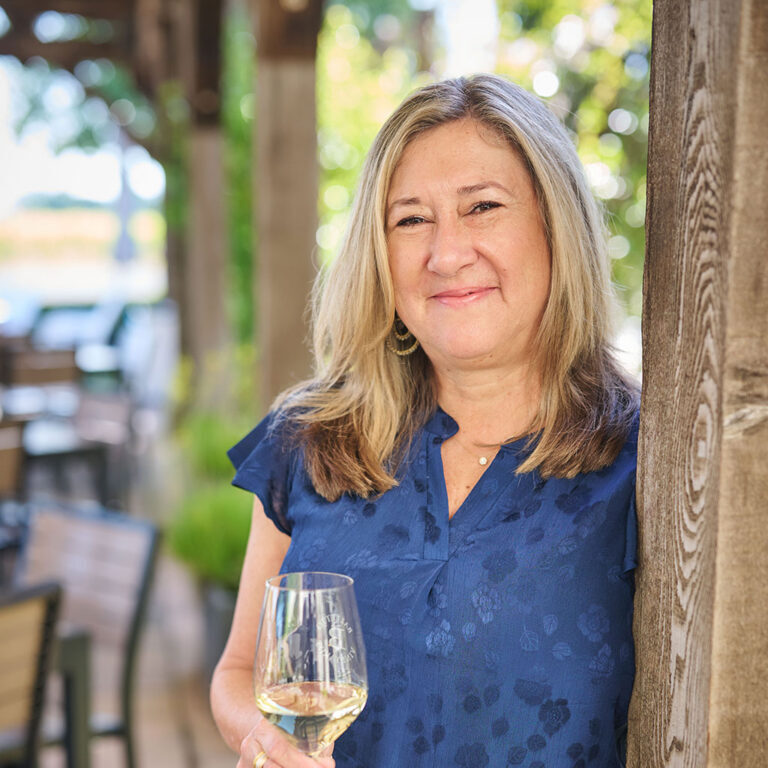 Smiling woman with long blonde hair holding a glass of white wine, standing outdoors by a wooden post.