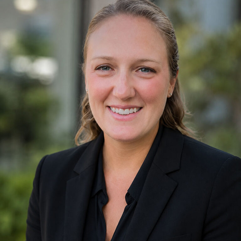 Woman in a black blazer smiling outdoors with greenery and a blurred building in the background.