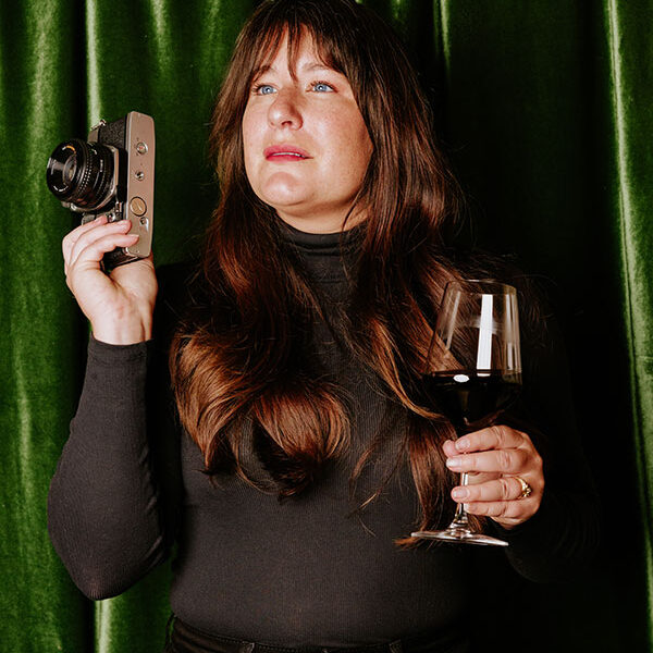 Woman in black holding a vintage camera and a glass of red wine, seated in front of green velvet curtains.