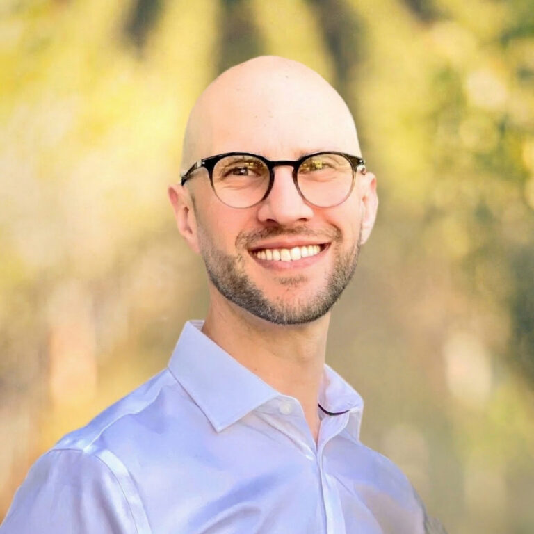 Bald man with glasses smiling, wearing a white shirt, standing outdoors with a blurred green background.