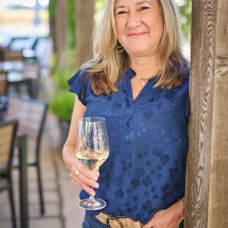 Woman with blonde hair in blue blouse holding a glass of white wine, smiling outdoors by a wooden post.
