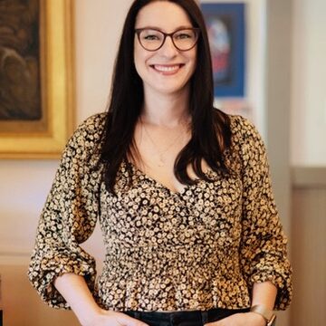 Woman with long dark hair, glasses, and a floral blouse smiling with hands in her pockets indoors.