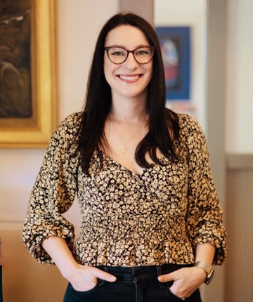 Woman with long dark hair, glasses, and a floral blouse smiling with hands in her pockets indoors.
