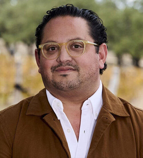 A man with short dark hair, glasses, and a brown jacket over a white shirt, outdoors in a blurred background.