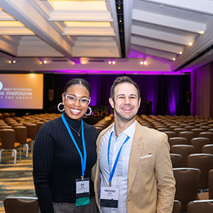 Two people wearing name tags and lanyards smile in a large, empty conference room with rows of chairs, excited about the upcoming event.