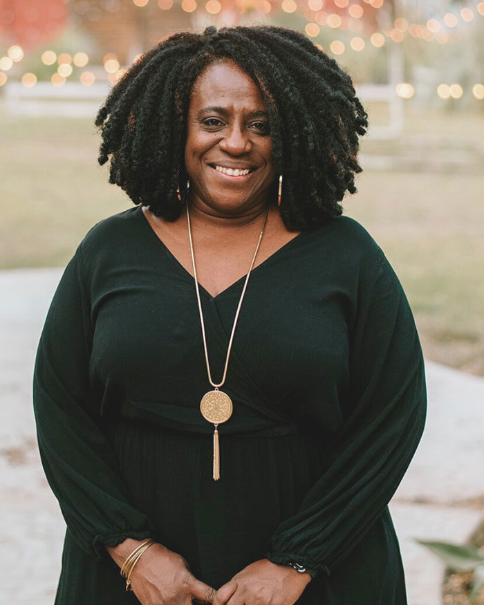 Smiling woman with natural hair in a black dress and gold necklace, standing outdoors with blurred lights behind her.