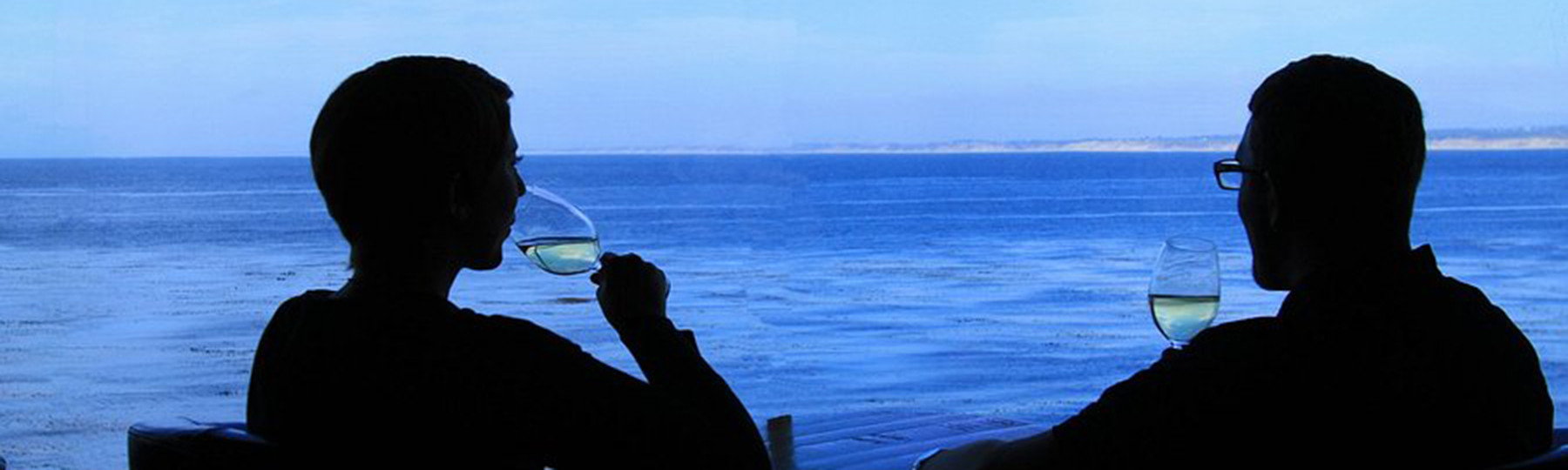 Two people with wine glasses sit facing the ocean, enjoying the view through a large window.