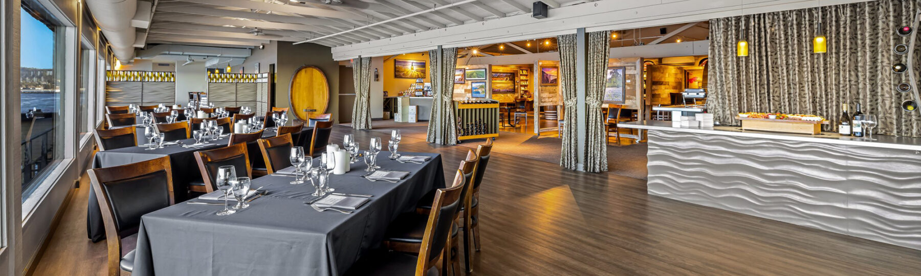 Spacious modern restaurant with large windows, set tables, wooden chairs, and a wavy-front bar area.