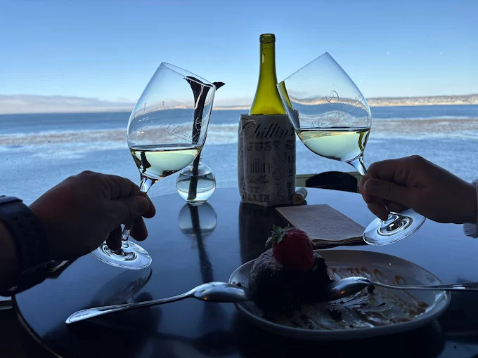 Two people clinking glasses of white wine over a plate with a chocolate dessert overlooking the ocean.