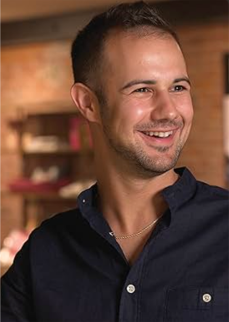 Smiling man with short hair and a trimmed beard, wearing a dark shirt, in a warmly lit indoor setting.